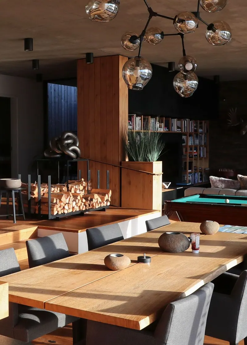 Dining room with a long teak table, grey chairs and a glass globe chandelier, a stacked log wall behind