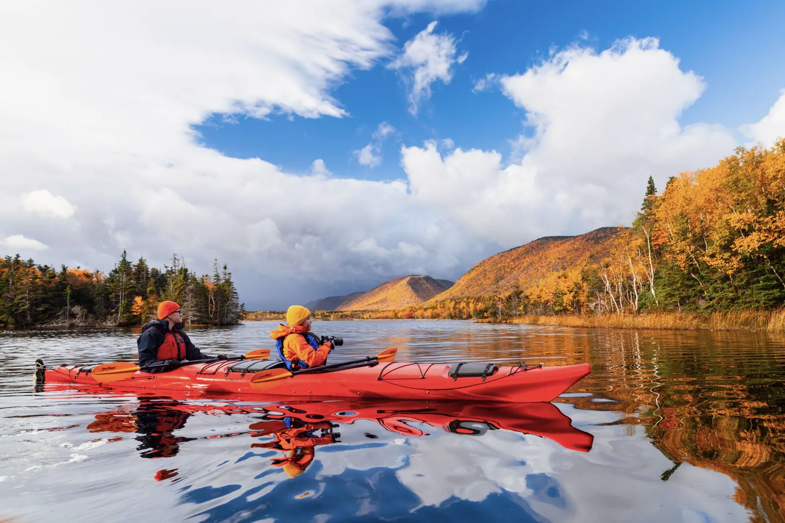Two paddlers in a red kayak on a still river below autumn-coloured hills