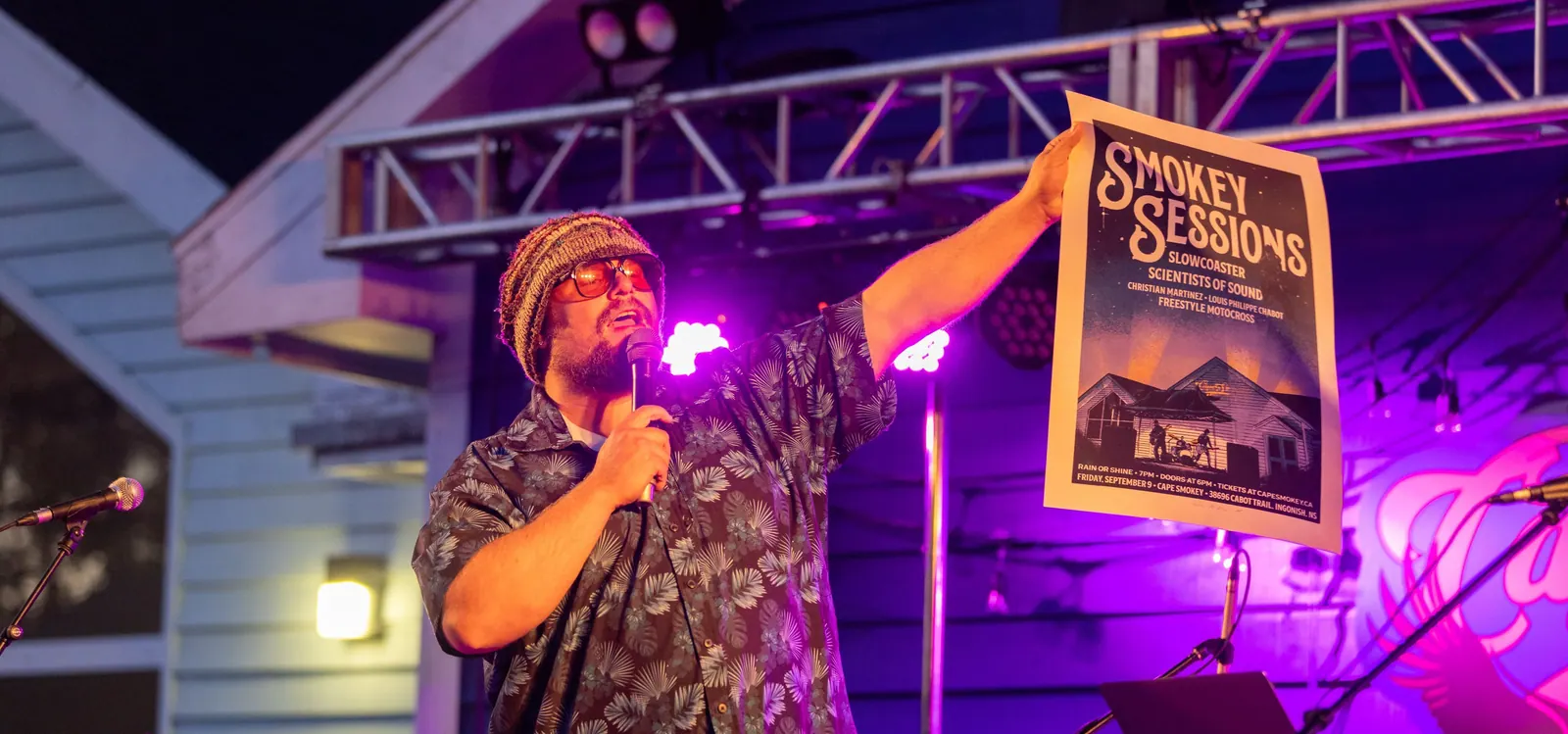 Musician singing on an outdoor stage at a Smokey Sessions concert, holding up the event poster