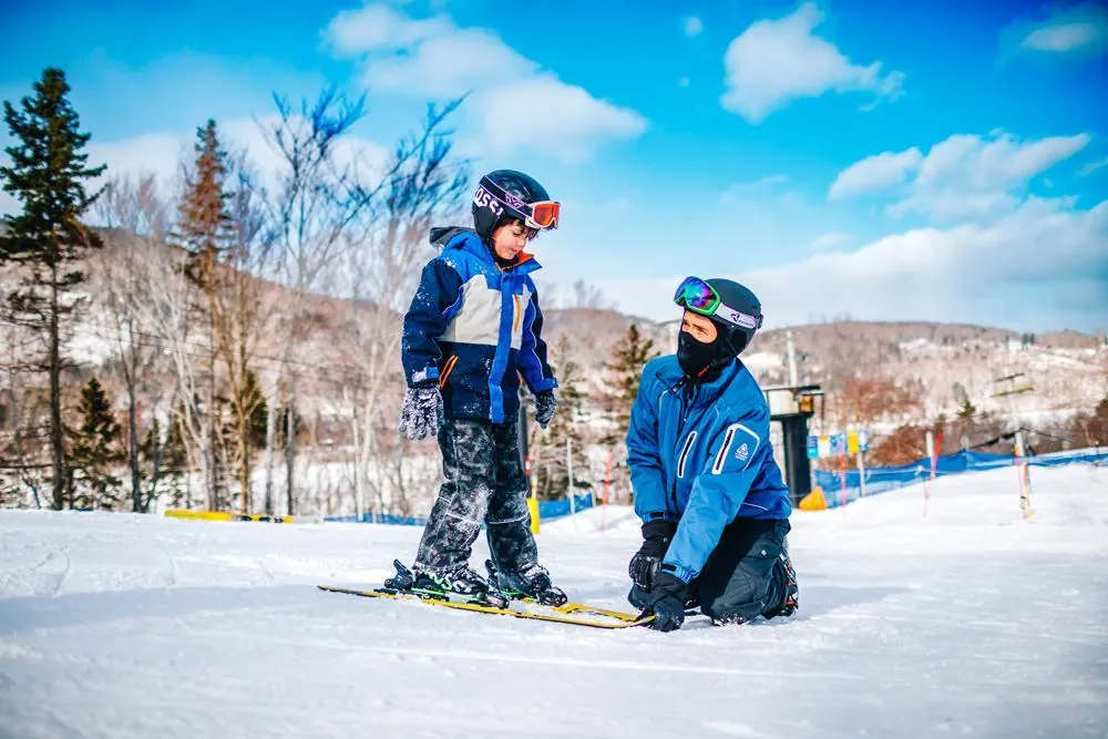 Snow School instructor kneeling to help a young skier on the beginner slope