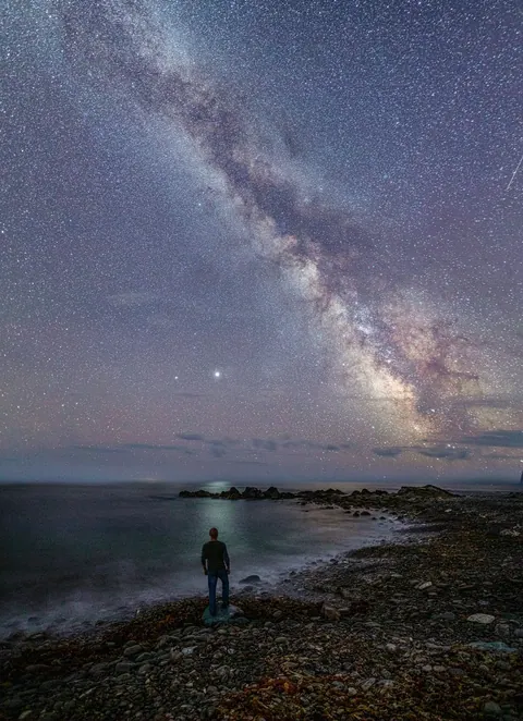 The Milky Way above a person standing on a rocky shore at night