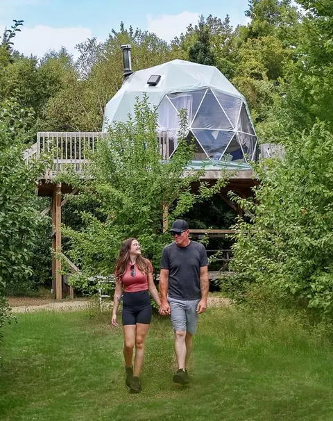 Couple walking past a glamping dome among green trees at Cabot Shores