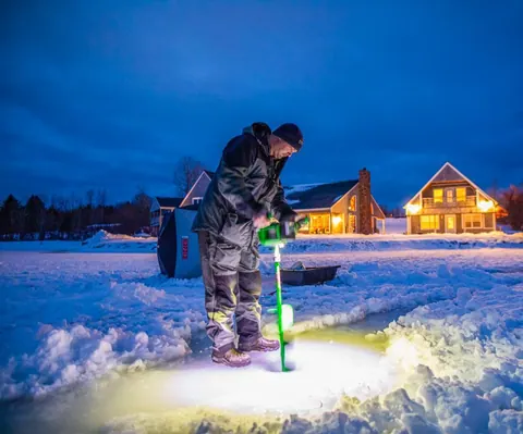 Angler drilling a hole through lake ice at dusk