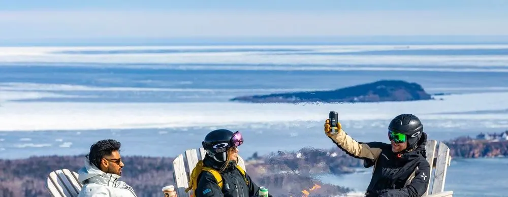 Friends in winter gear relaxing in Adirondack chairs at the summit, raising a drink above the ocean