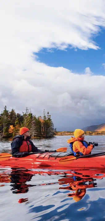 Two paddlers in a red kayak on a still river below autumn-coloured hills