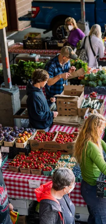 Shoppers at a farmer’s market stall with fresh strawberries, plums and vegetables