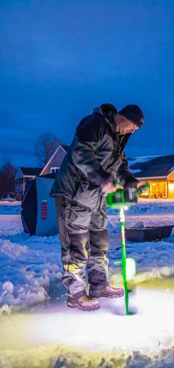 Angler drilling a hole in the lake ice at dusk, lit cottages behind