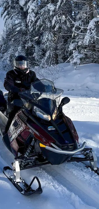 Two snowmobilers riding a groomed trail through snow-laden spruce forest