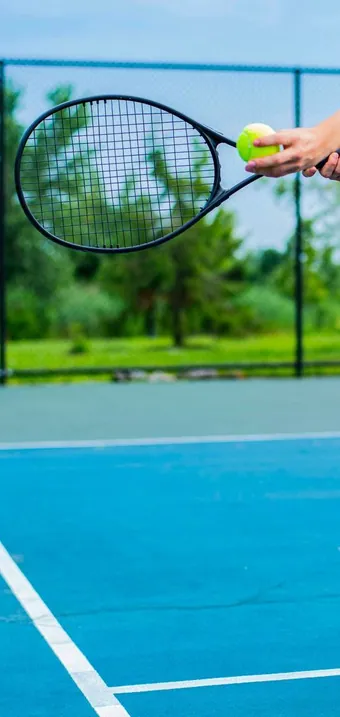 Tennis racket raised above a blue outdoor court lined with trees