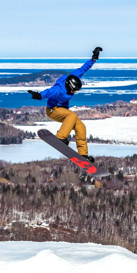 Snowboarder jumping above snowy trees with the ocean behind