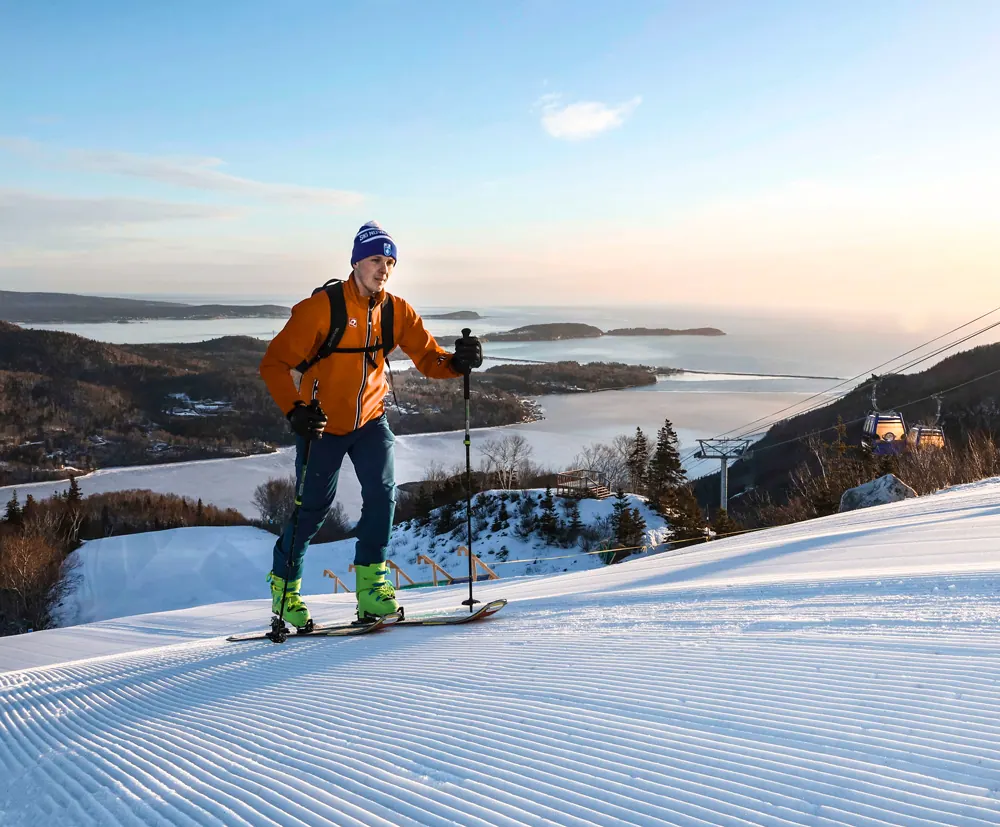 Ski tourer skinning up a groomed slope with the bay and gondola behind