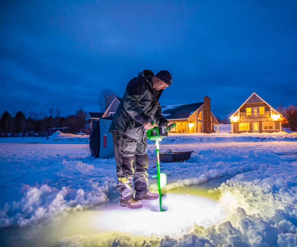 Angler drilling a hole in the lake ice at dusk, lit cottages behind